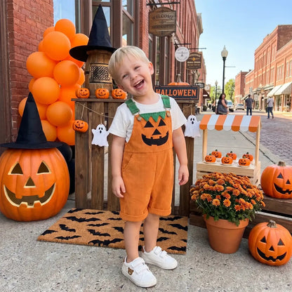 A young toddler boy wearing orange corduroy overalls with a green and orange pumpkin face, standing in front of a Halloween-themed storefront decorated with pumpkins, balloons, and mums.