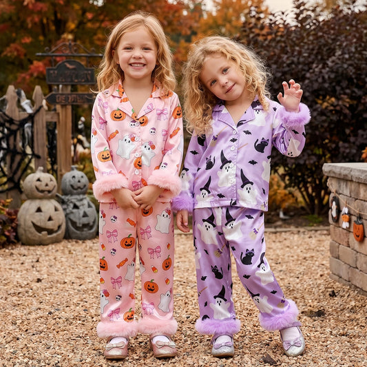 Two young girls wearing matching Halloween-themed pajama sets, standing outdoors in a garden setting. One set is light pink with orange jack-o'-lanterns, white ghosts, and purple bows with fluffy trim.