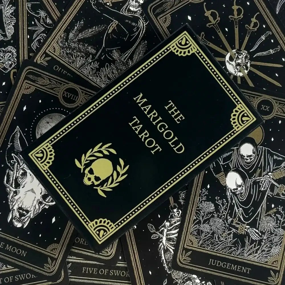 The Marigold Tarot deck's black card box with gold foil detailing is displayed diagonally on top of several tarot cards, including the Eight of Swords, Five of Swords, and Judgment cards, all featuring a matching black and gold aesthetic.
