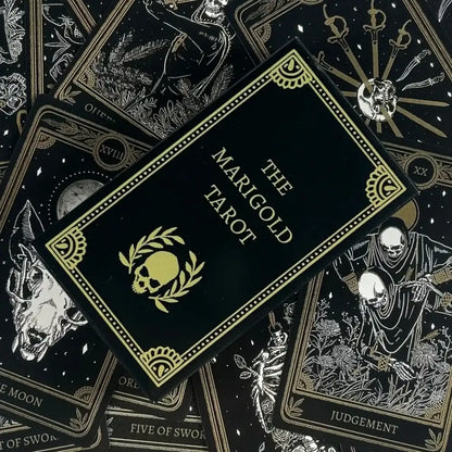 The Marigold Tarot deck's black card box with gold foil detailing is displayed diagonally on top of several tarot cards, including the Eight of Swords, Five of Swords, and Judgment cards, all featuring a matching black and gold aesthetic.