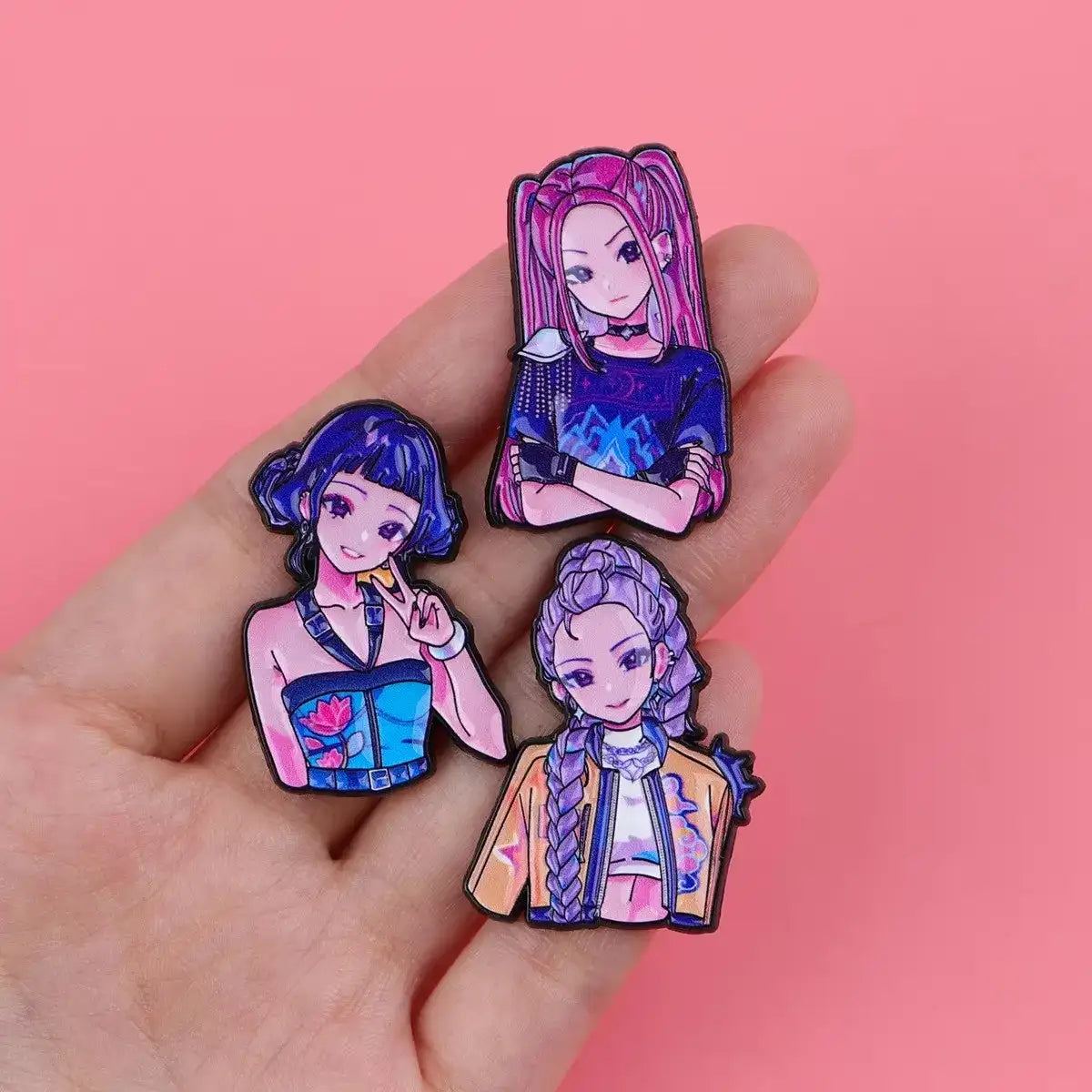 Three colorful anime-style enamel pins held on a person's fingers against a solid pink background.