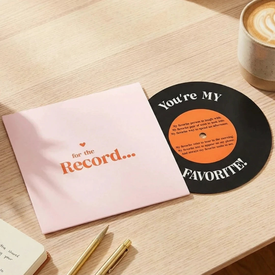 Vinyl record with 'You're My Favorite!' text on a pink card, next to a cup of coffee on a wooden surface.