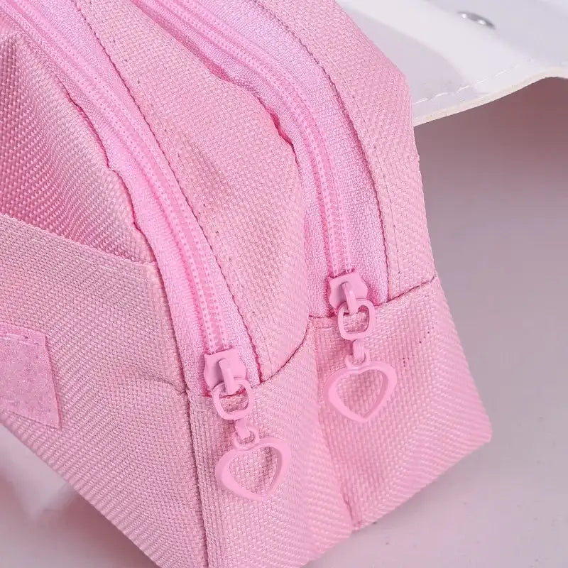 A close-up of a light pink canvas zippered pouch with matching pink zippers and heart-shaped zipper pulls, shown on a pale pink background.