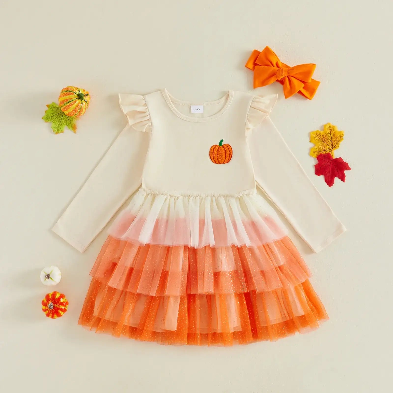 A toddler girl's long-sleeve dress with a cream bodice, a small orange pumpkin graphic on the chest, and a tiered tulle skirt in cream, light pink, and orange. The dress is styled in a flat-lay with autumn props including a matching orange bow, a small pumpkin, and fall leaves on a light beige background.