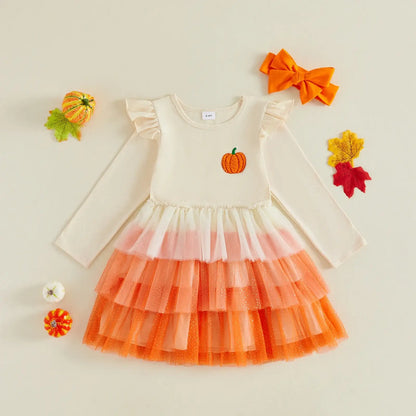 A toddler girl's long-sleeve dress with a cream bodice, a small orange pumpkin graphic on the chest, and a tiered tulle skirt in cream, light pink, and orange. The dress is styled in a flat-lay with autumn props including a matching orange bow, a small pumpkin, and fall leaves on a light beige background.