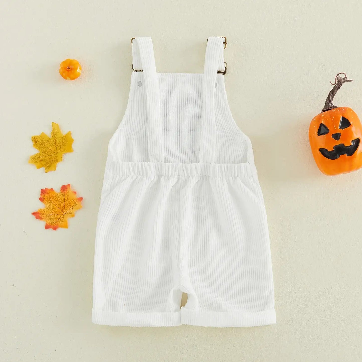 A white ribbed short-sleeve baby romper with shoulder straps, laid flat on a light beige background with small autumn props including a pumpkin, maple leaves, and a tiny orange object.