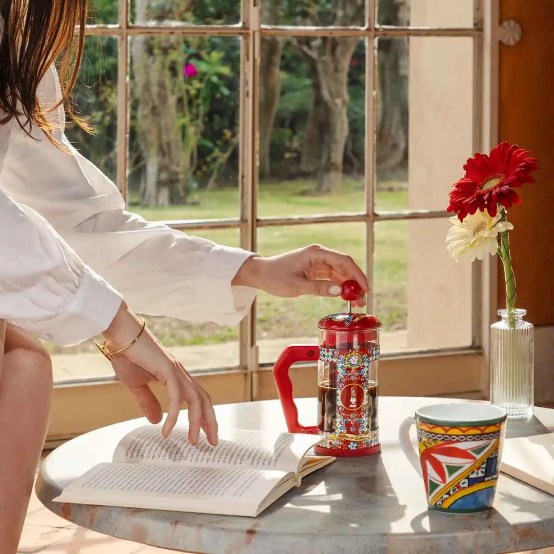 Person making coffee using a French press on a table with a book and colorful mug, near a window with a garden view.