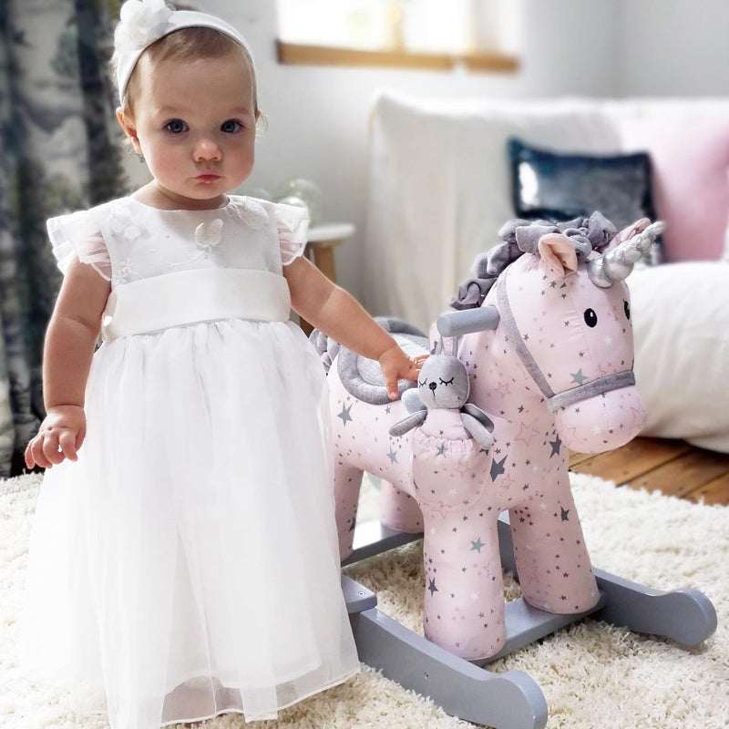 Baby in a white dress standing next to a pink rocking unicorn toy in a room.