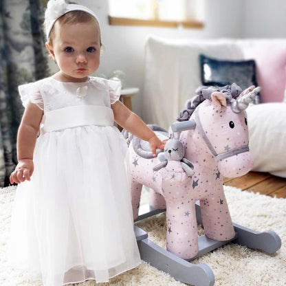 Baby in a white dress standing next to a pink rocking unicorn toy in a room.