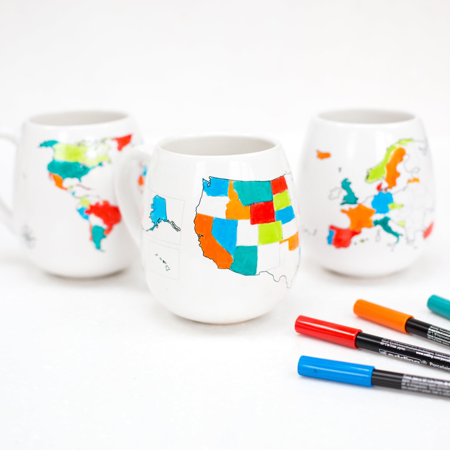 Three white ceramic cups with colorful map designs on a white background, accompanied by markers.