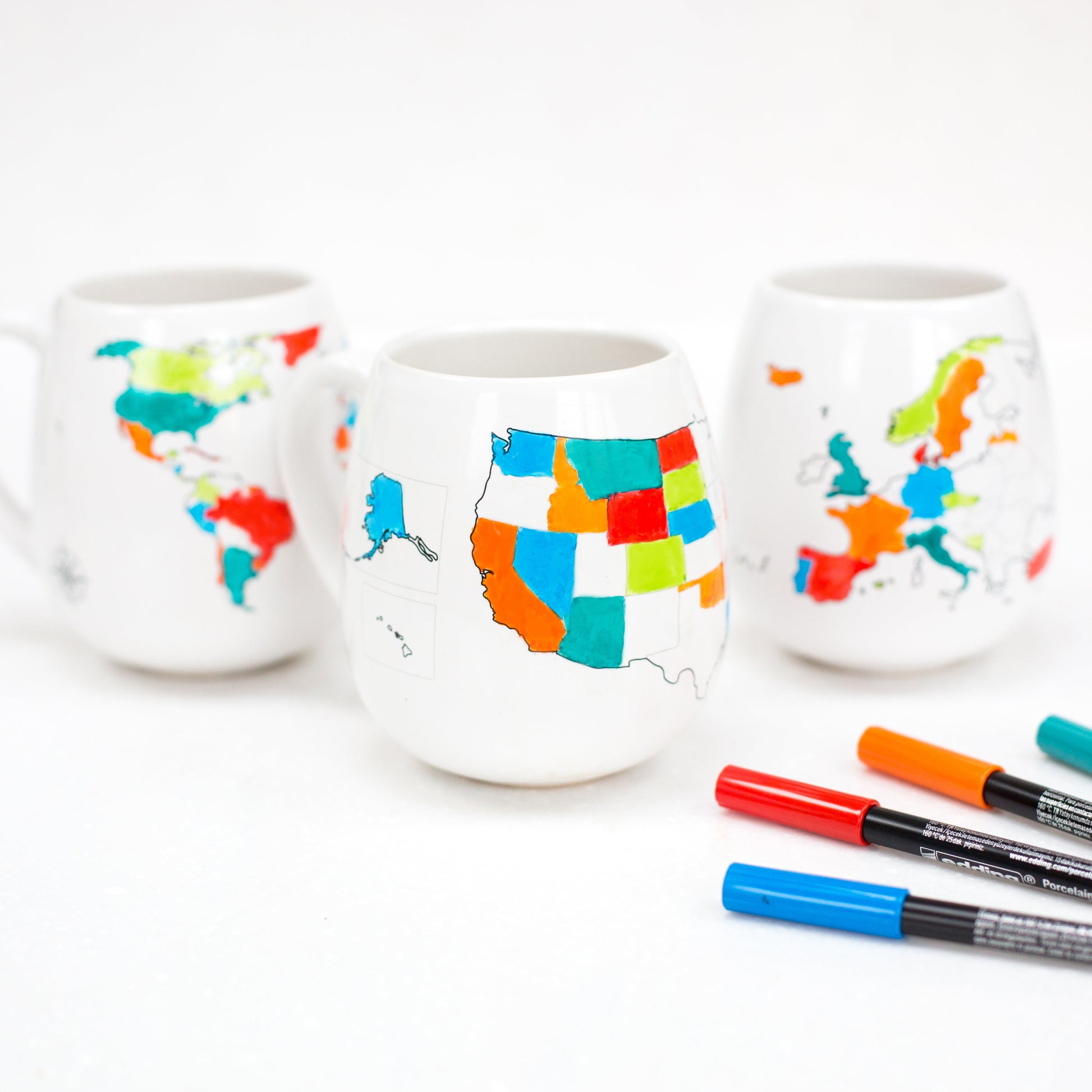 Three white ceramic cups with colorful map designs on a white background, accompanied by markers.
