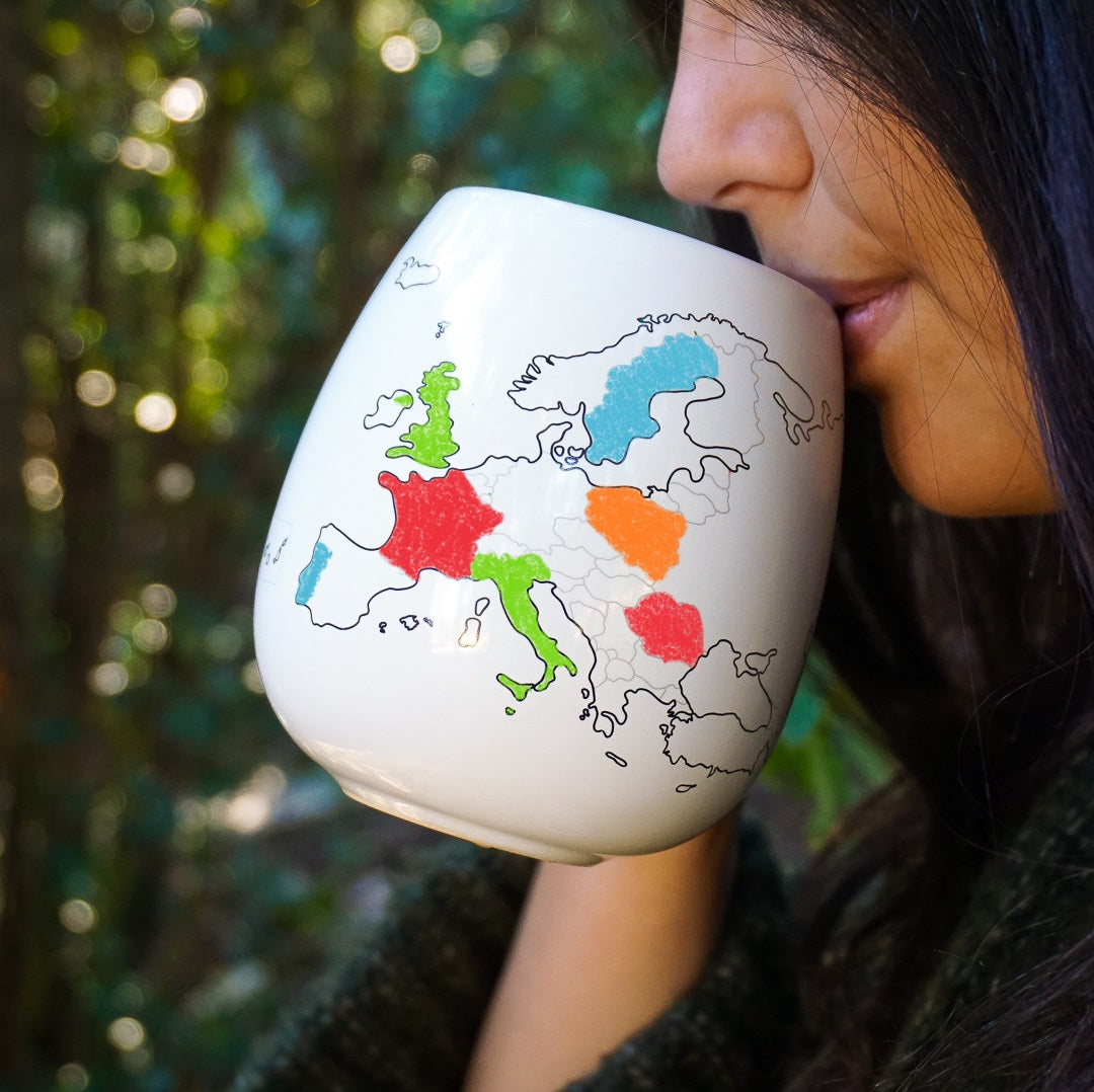 Person holding a mug with a map of Europe in colorful markers against a blurred green background