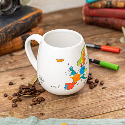 White mug with a colorful map design on a wooden surface with coffee beans and books.