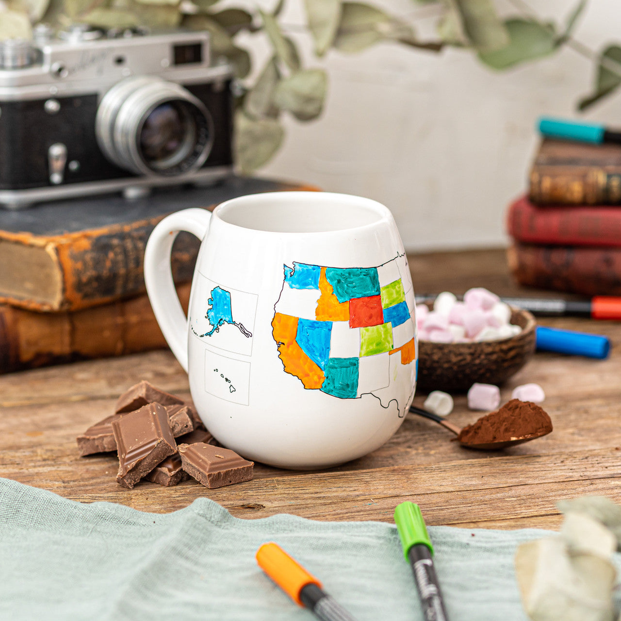 White mug with colorful map design on a wooden table with chocolate, books, and a camera in the background.
