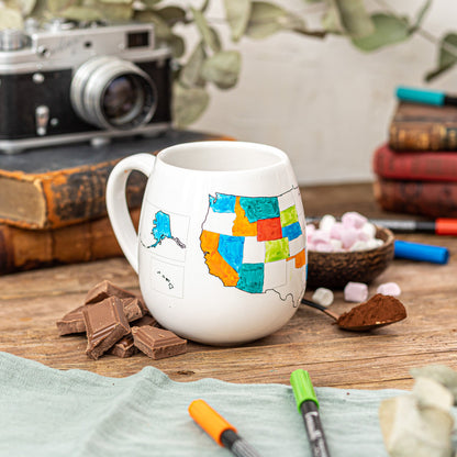 White mug with colorful map design on a wooden table with chocolate, books, and a camera in the background.