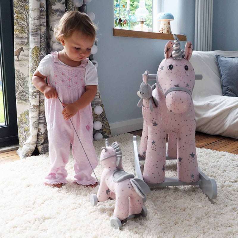 Child playing with a pink rocking horse toy in a room with a window and furniture.