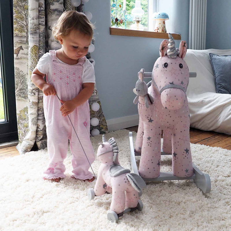 Child playing with a pink rocking horse toy in a room with a window and furniture.