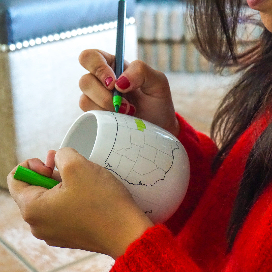Person coloring a white mug with green markers indoors.