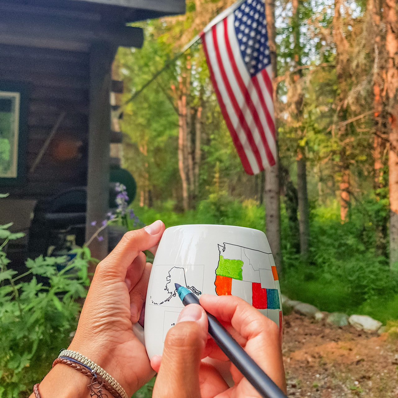 Person coloring a map on a ceramic mug with an American flag and cabin in the background