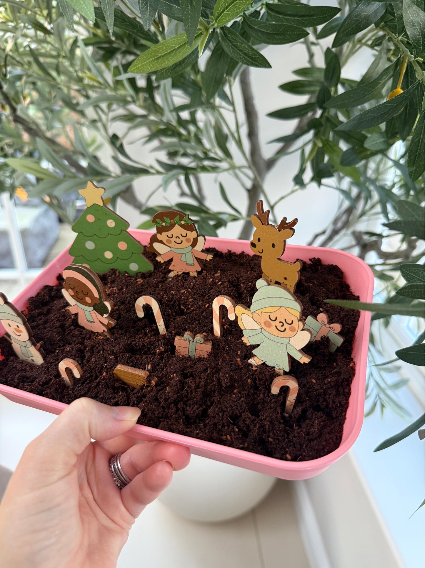 Pink tray with soil and miniature fairy garden figures, including trees and animals, held by a hand.