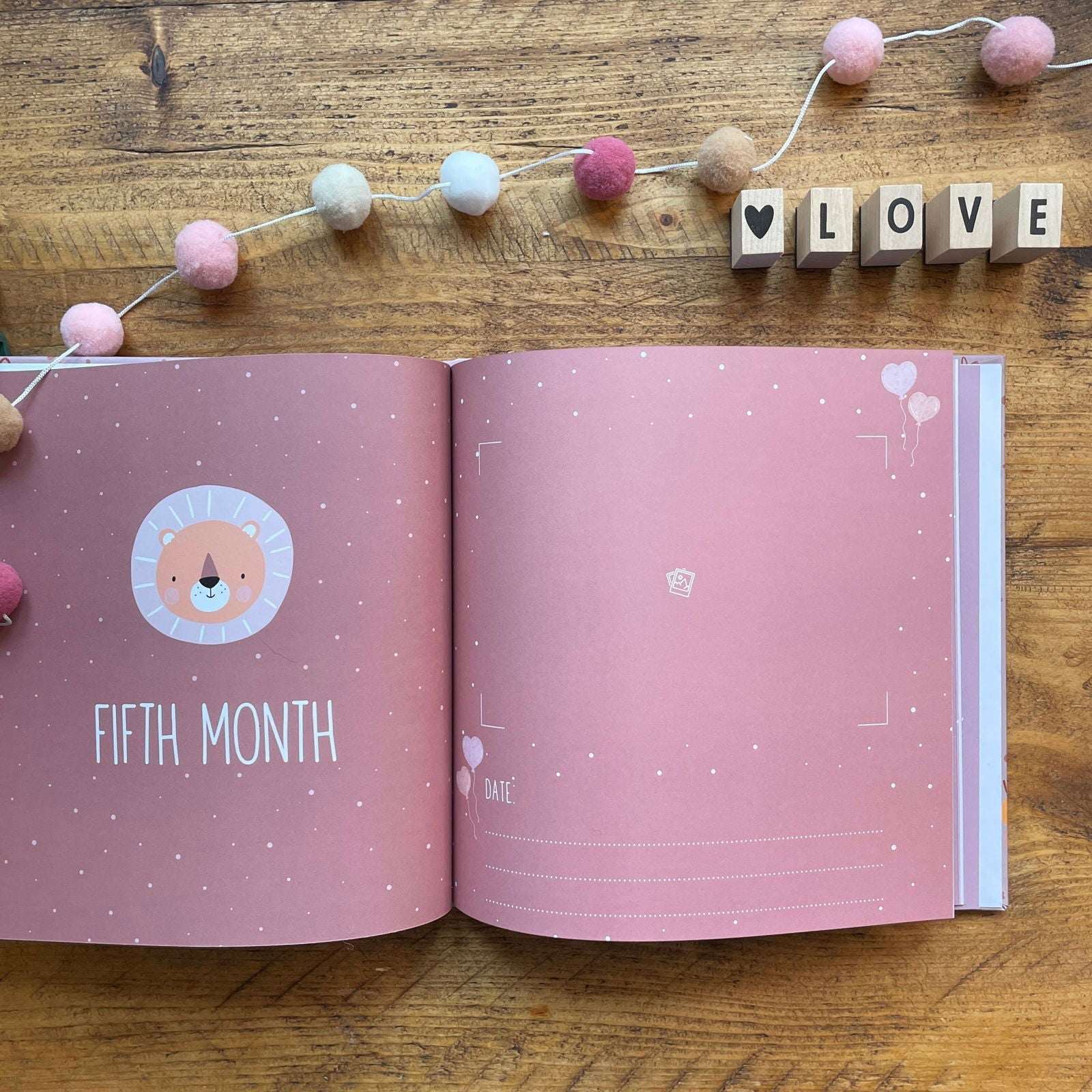 Open book with a pink page showing 'Fifth Month' on a wooden surface with decorative elements.