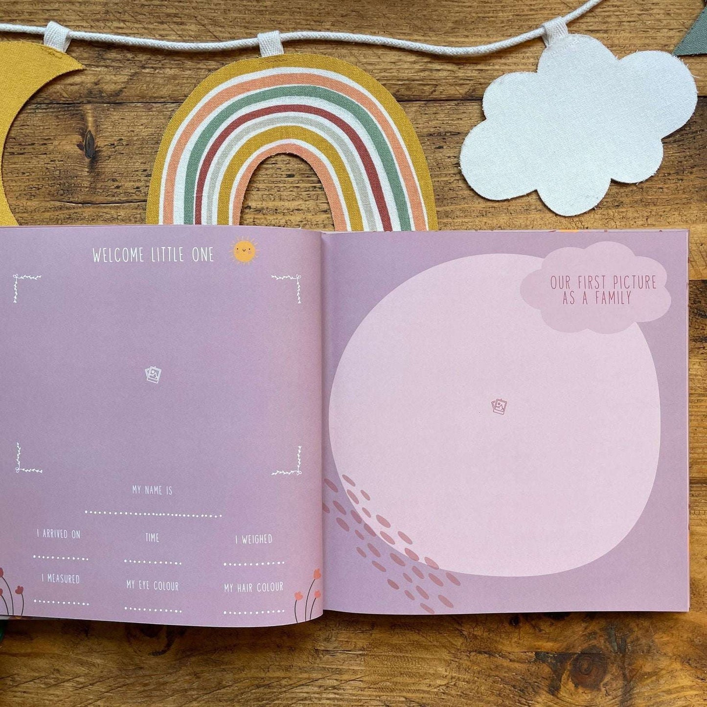 Open book with baby milestone tracker on a wooden surface with rainbow and cloud decorations.