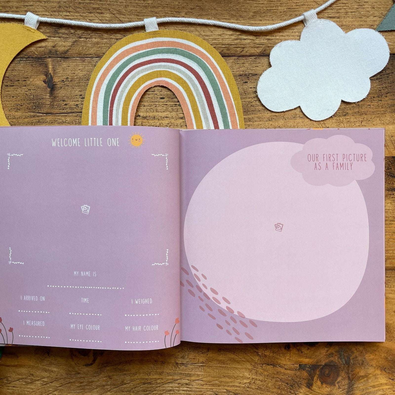 Open book with baby milestone tracker on a wooden surface with rainbow and cloud decorations.