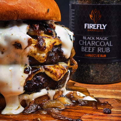 Stacked burger with cheese and onions next to a bottle of Firefly Black Magic Charcoal Beef Rub on a wooden surface.