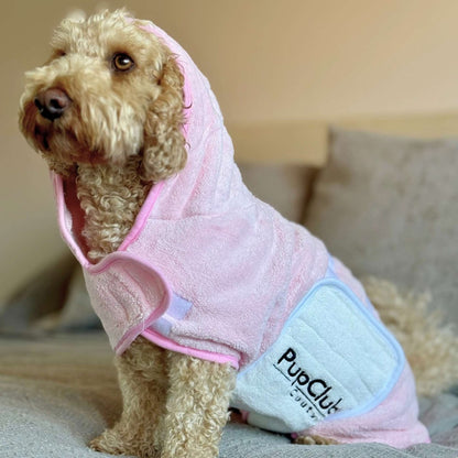 Dog wearing a pink hooded towel with 'PupClub' branding on a couch.