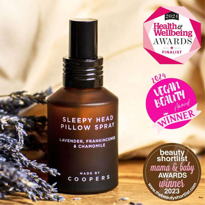 Sleepy Head Pillow Spray bottle with lavender, on a wooden surface, surrounded by lavender flowers, with awards logos displayed.