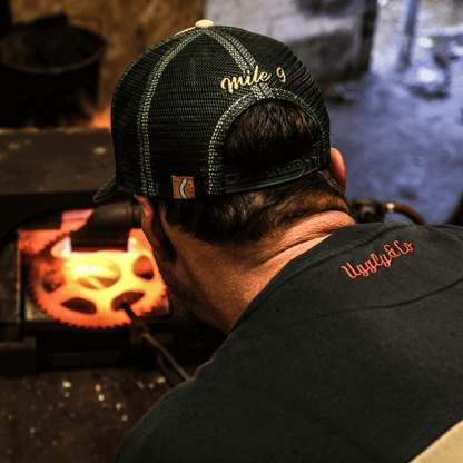 Person wearing a black cap and black shirt with red text, working on a glowing metal object.